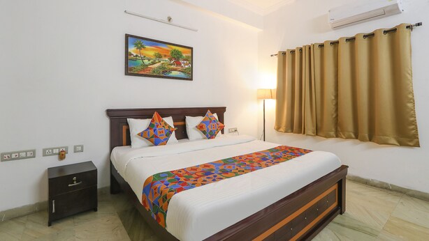 A neatly made bed with a colorful bedspread, a nightstand with a drawer, a wall-mounted air conditioner, and a painting above the bed.