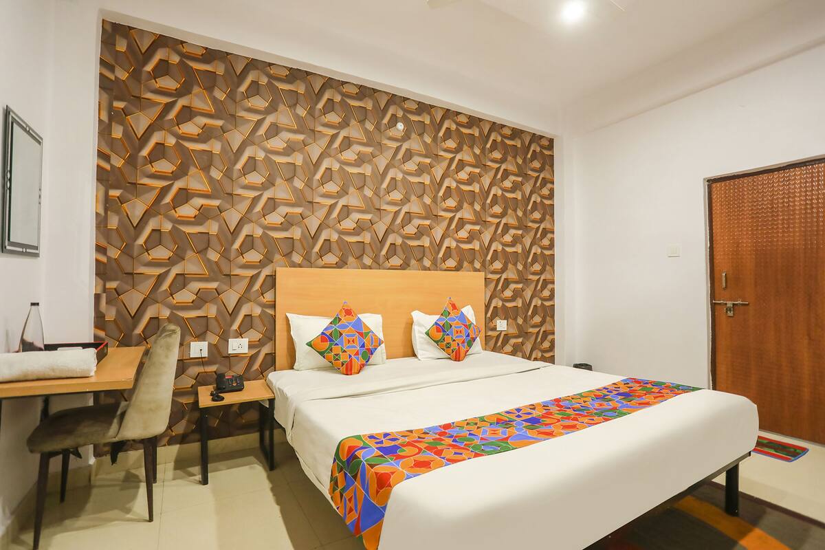 A hotel room with a geometric-patterned headboard, a bed with colorful bedding, a desk with a chair, and a wooden door.