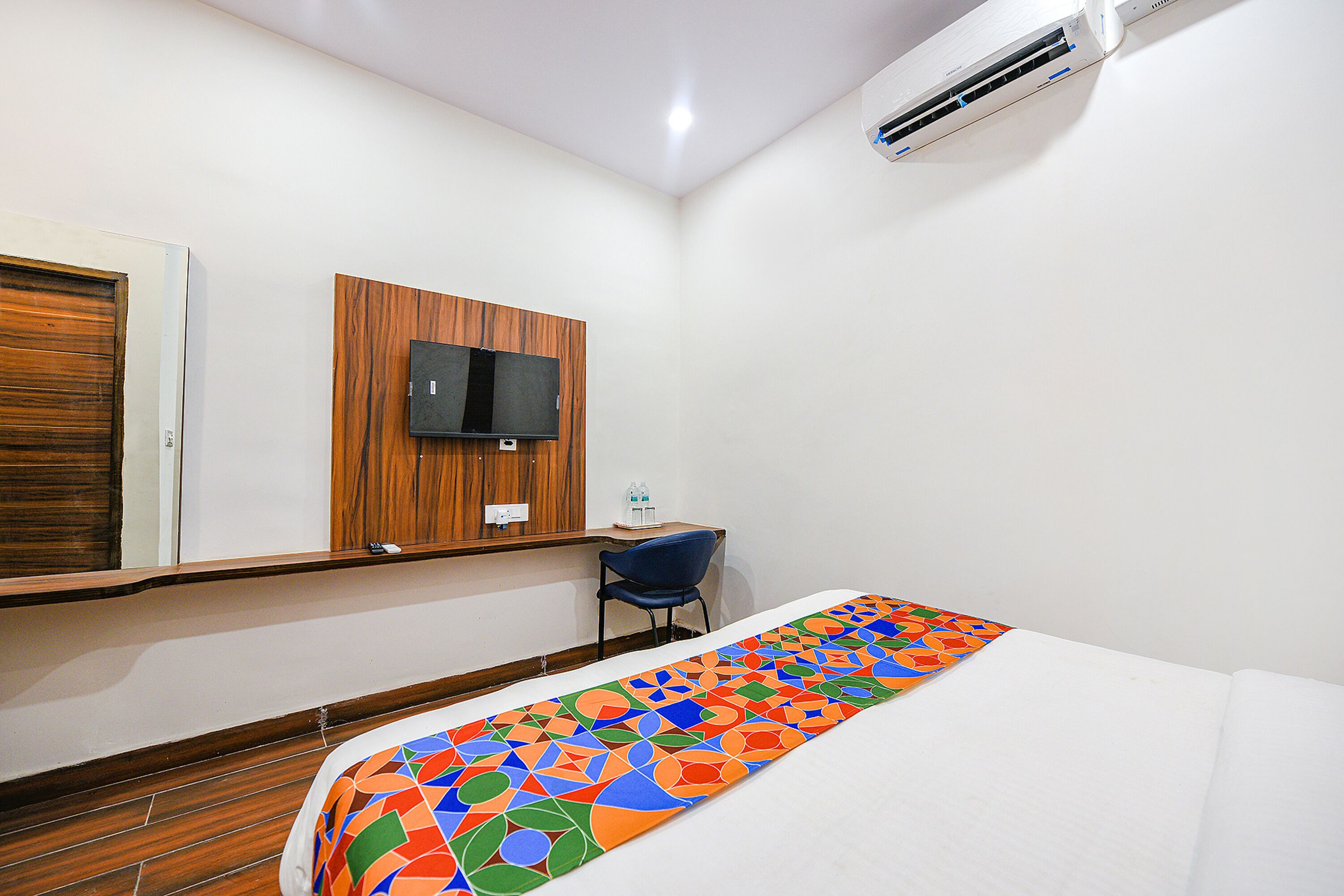 A hotel room with a bed, a desk, a chair, a TV mounted on the wall, and an air conditioning unit.