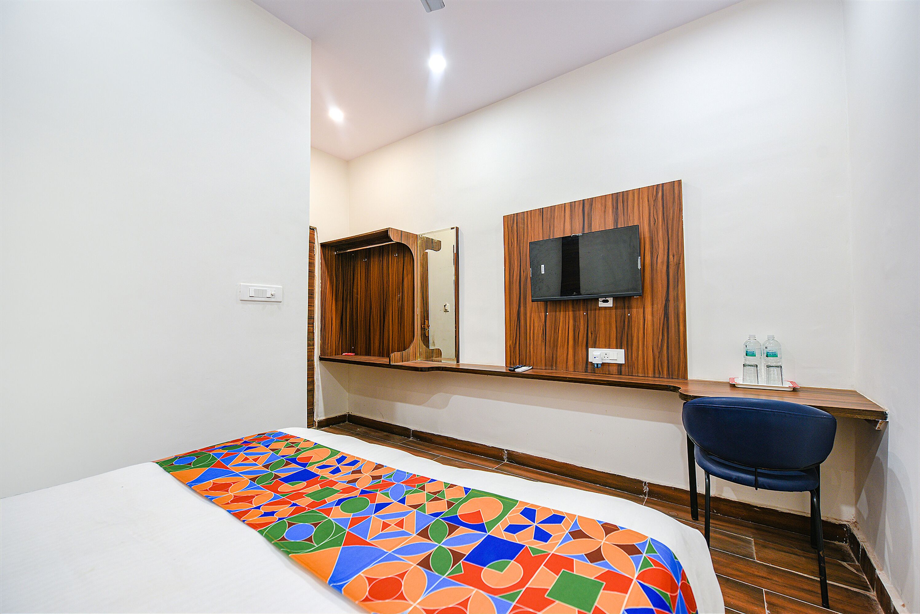 Quarto de hotel com cama, escrivaninha, cadeira, televisão fixada em um painel de madeira e espelho.