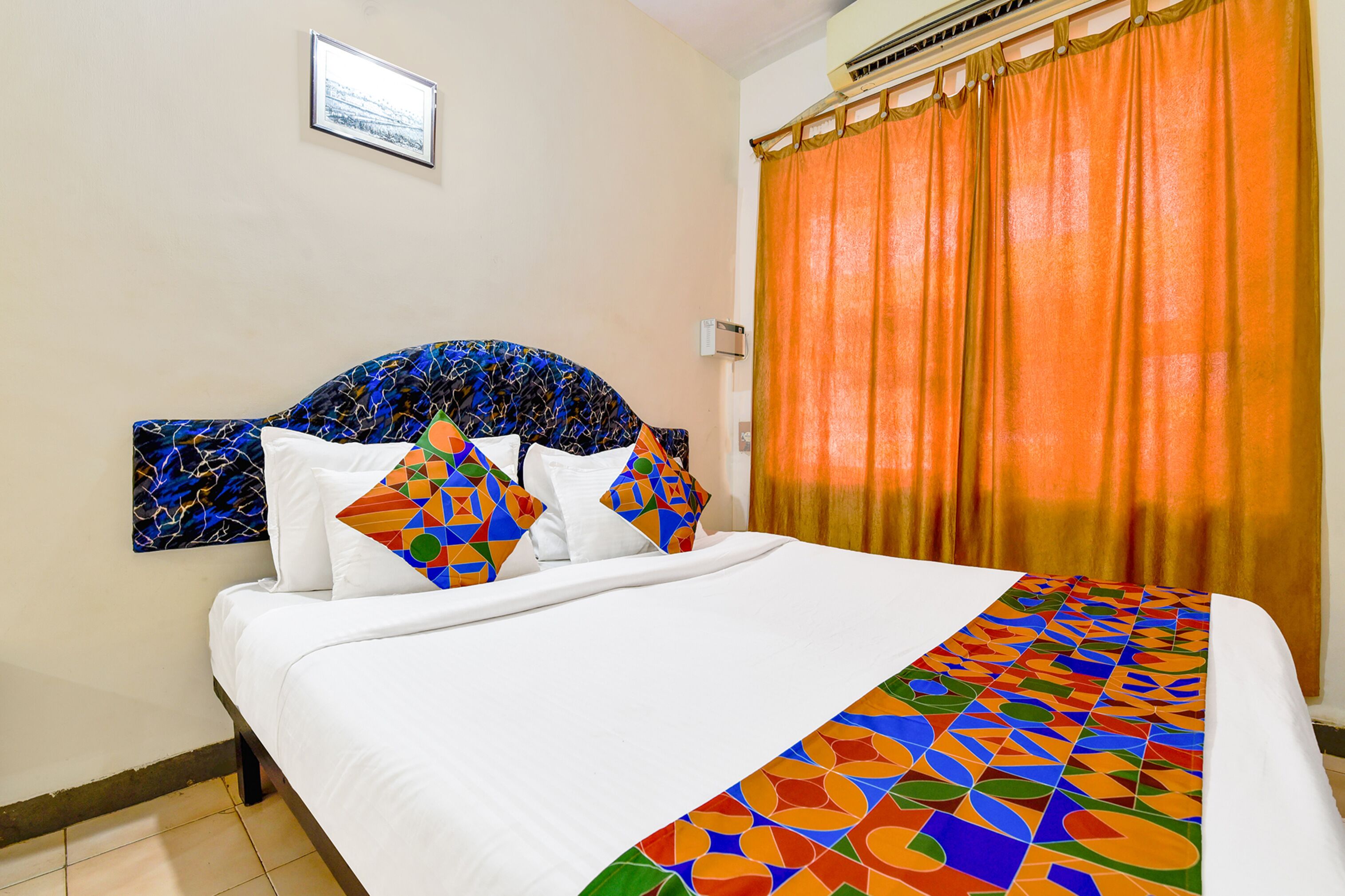 Ein Hotelzimmer mit einem Bett, einem Kopfstück mit geometrischem Muster, einer farbenfrohen Bettdecke und orangefarbenen Vorhängen.