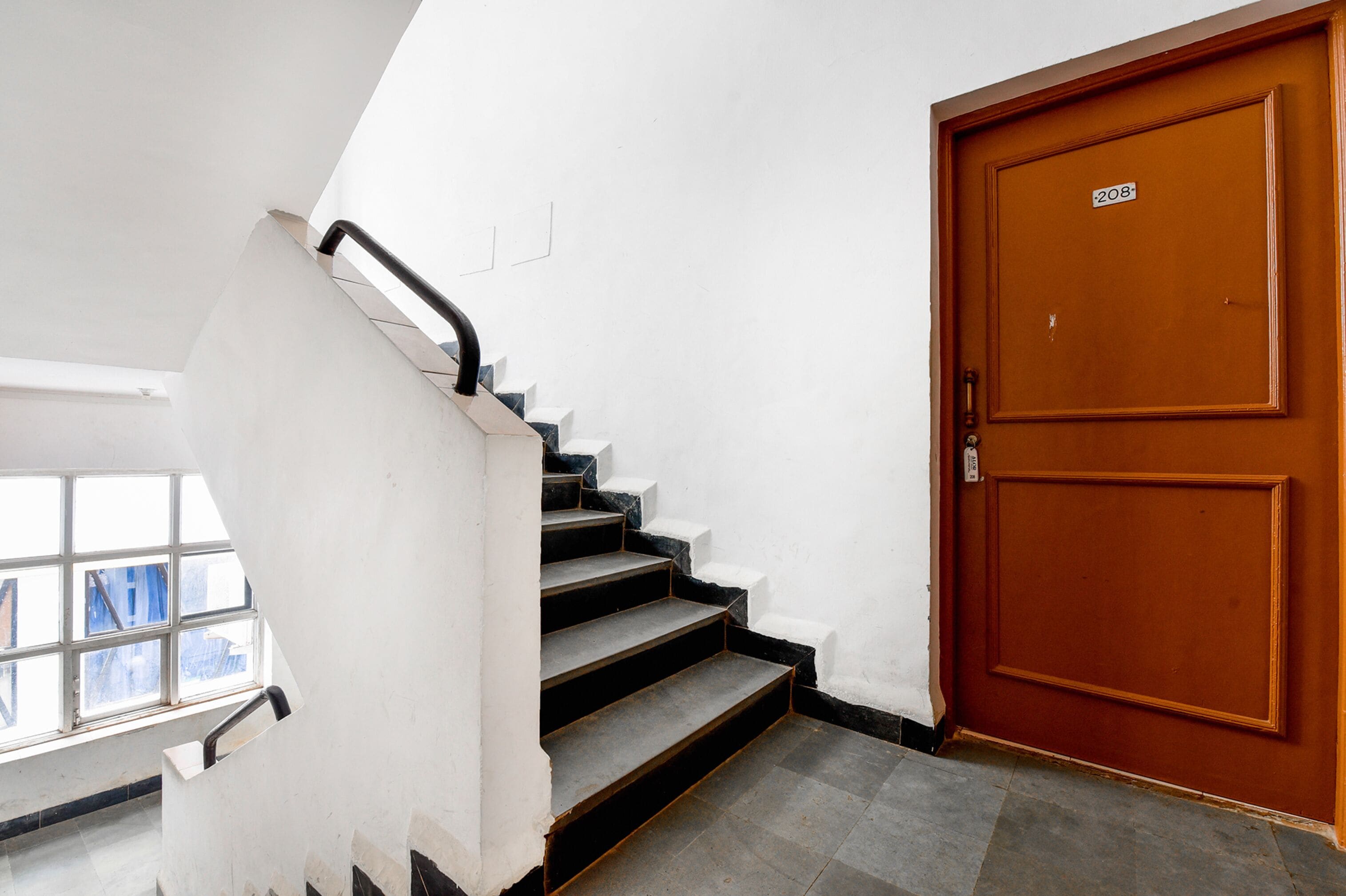 Un escalier avec une rampe, une porte en bois avec une plaque indiquant le numéro, et une fenêtre donnant sur l’extérieur.