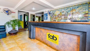 Zona de recepción de un hotel con piso de baldosas, un mostrador de recepción con el logo 'fab hotels', un ventilador grande y un mural que representa una escena costera.