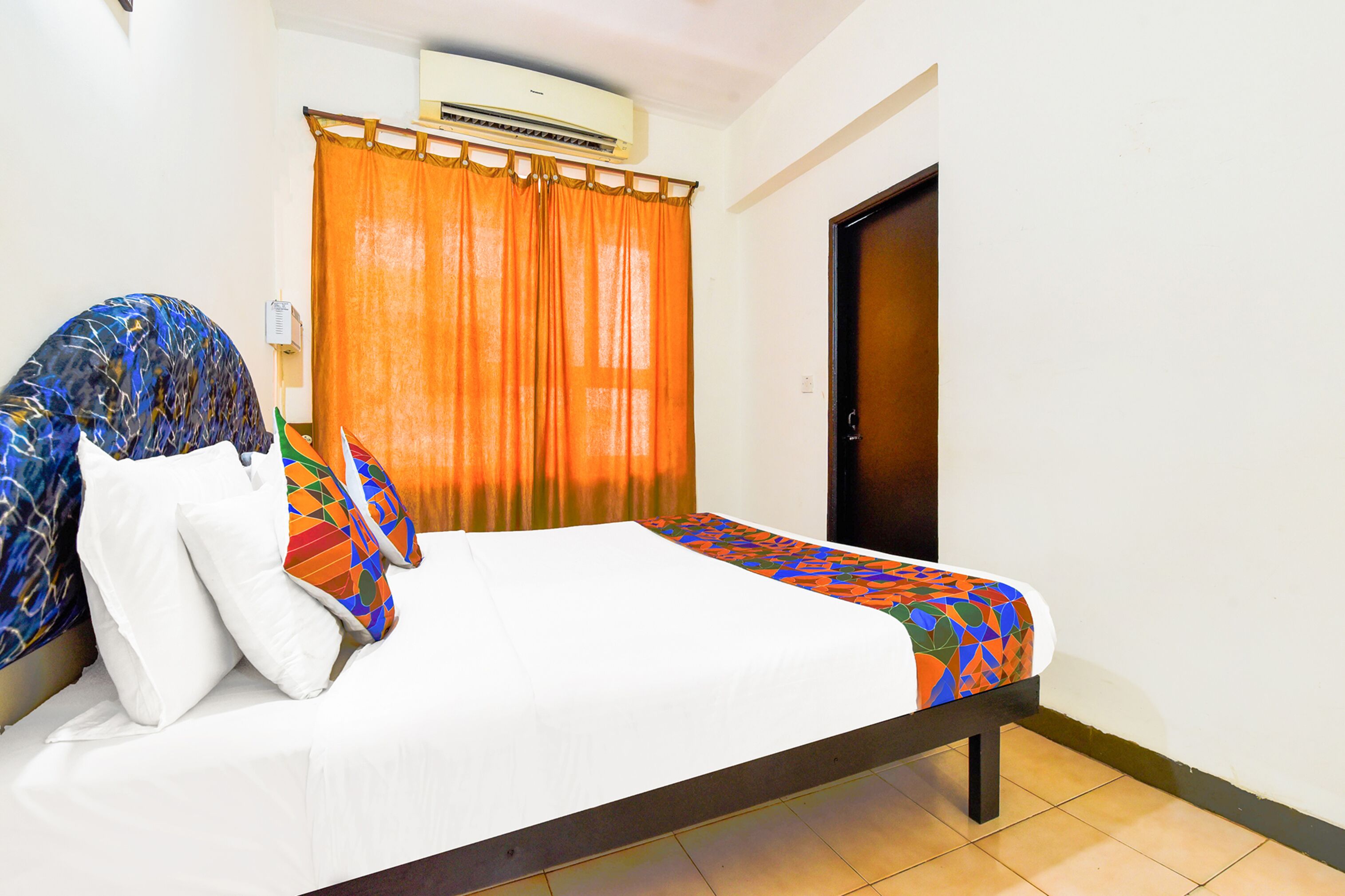 A hotel room with a bed, an air conditioner, an orange curtain, and a wooden door.
