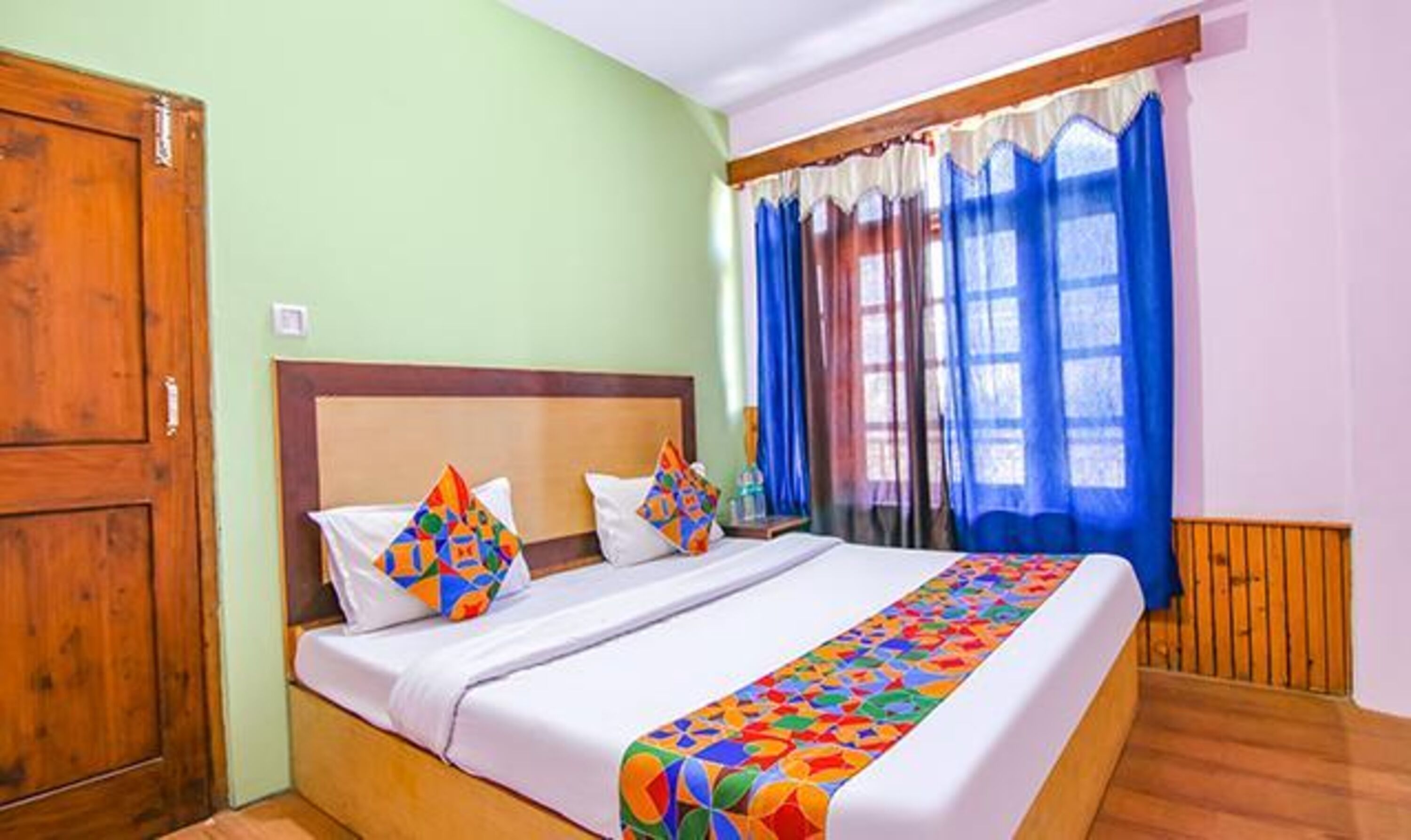 A neatly made bed with colorful pillows and a patterned bed runner, a wooden wardrobe, and a window with blue curtains.