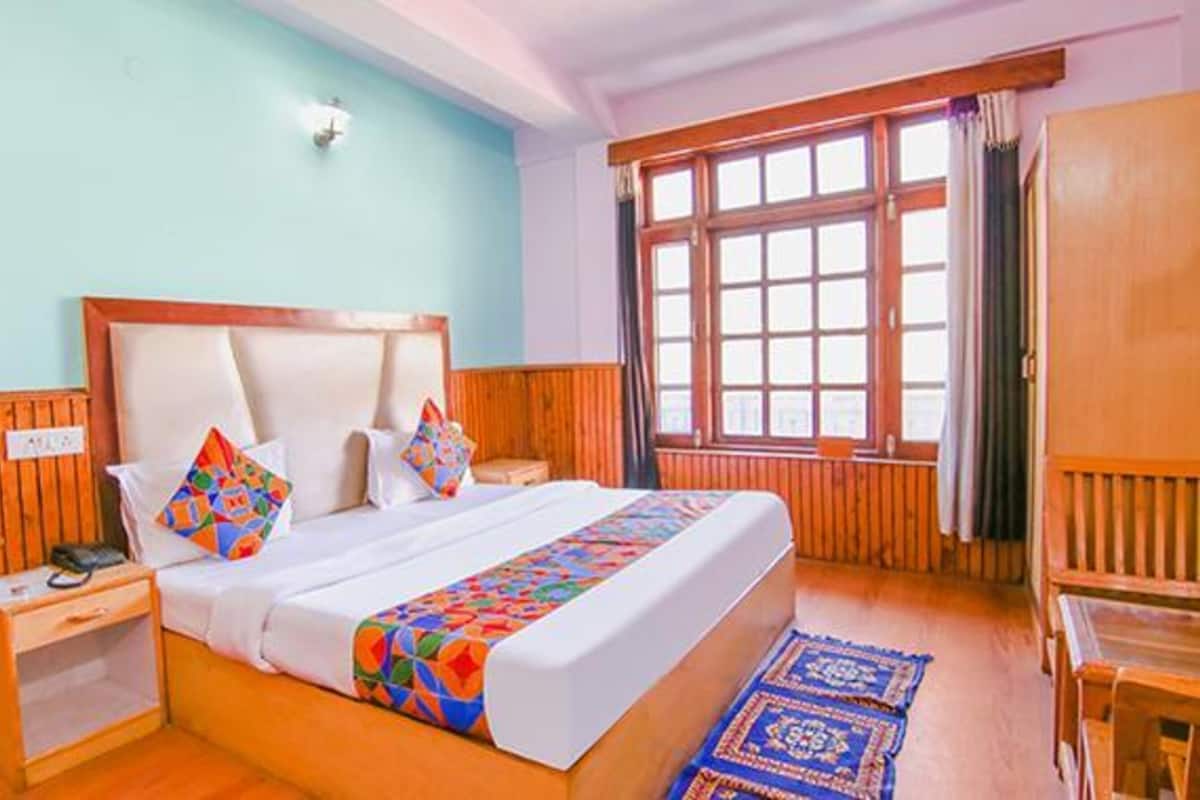 A bedroom with a wooden headboard, a bed with colorful bedding, a nightstand, a bedside table, a window with curtains, and a wooden bench.