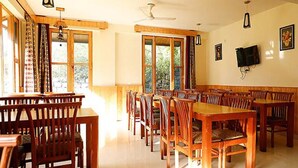 A dining area with wooden tables and chairs, large windows, and a ceiling fan. - Fabhotel Mahavir (Manali)