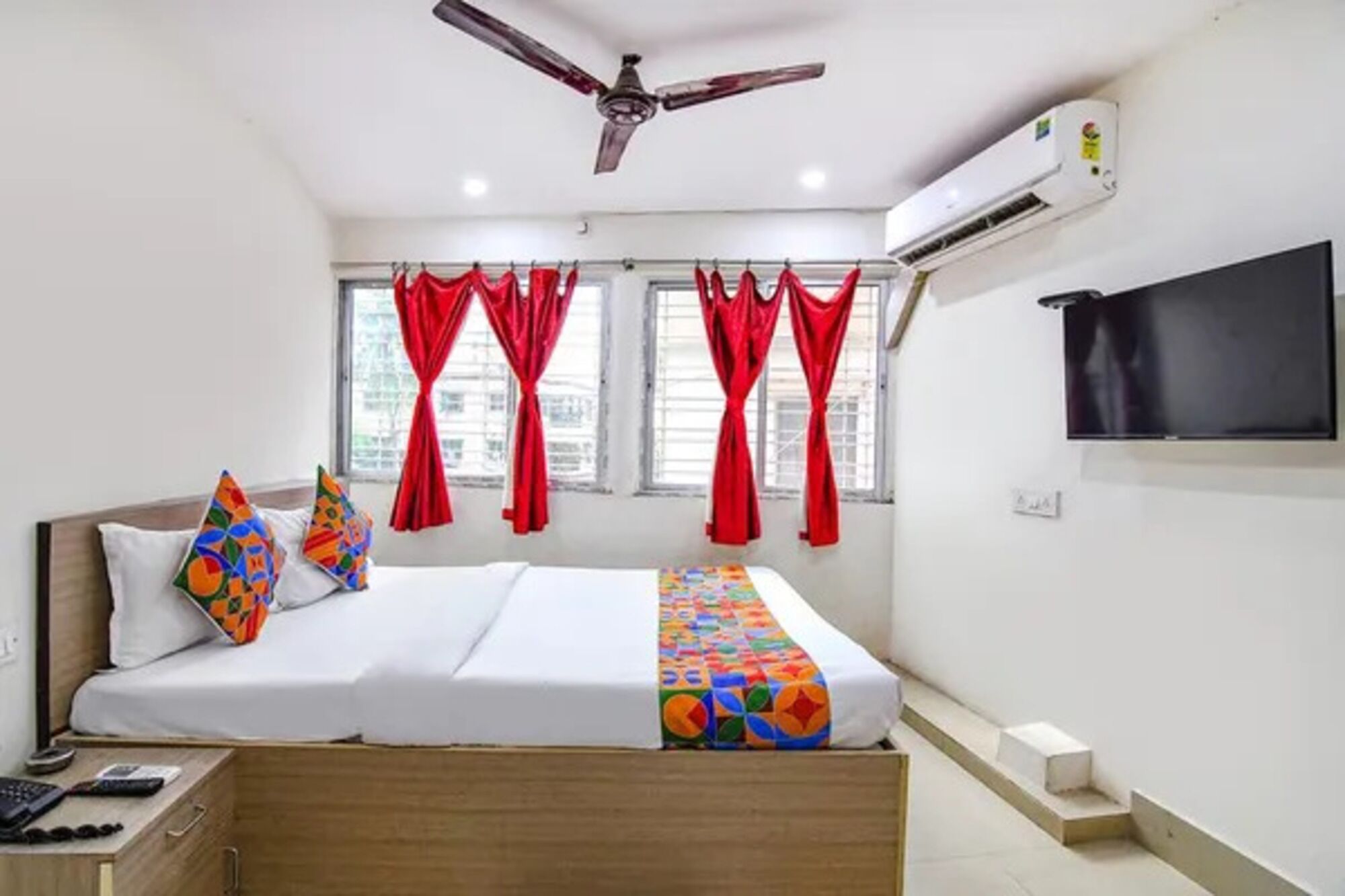 Habitación de hotel con una cama, ventilador de techo, equipo de aire acondicionado, televisor y ventana con cortinas rojas.