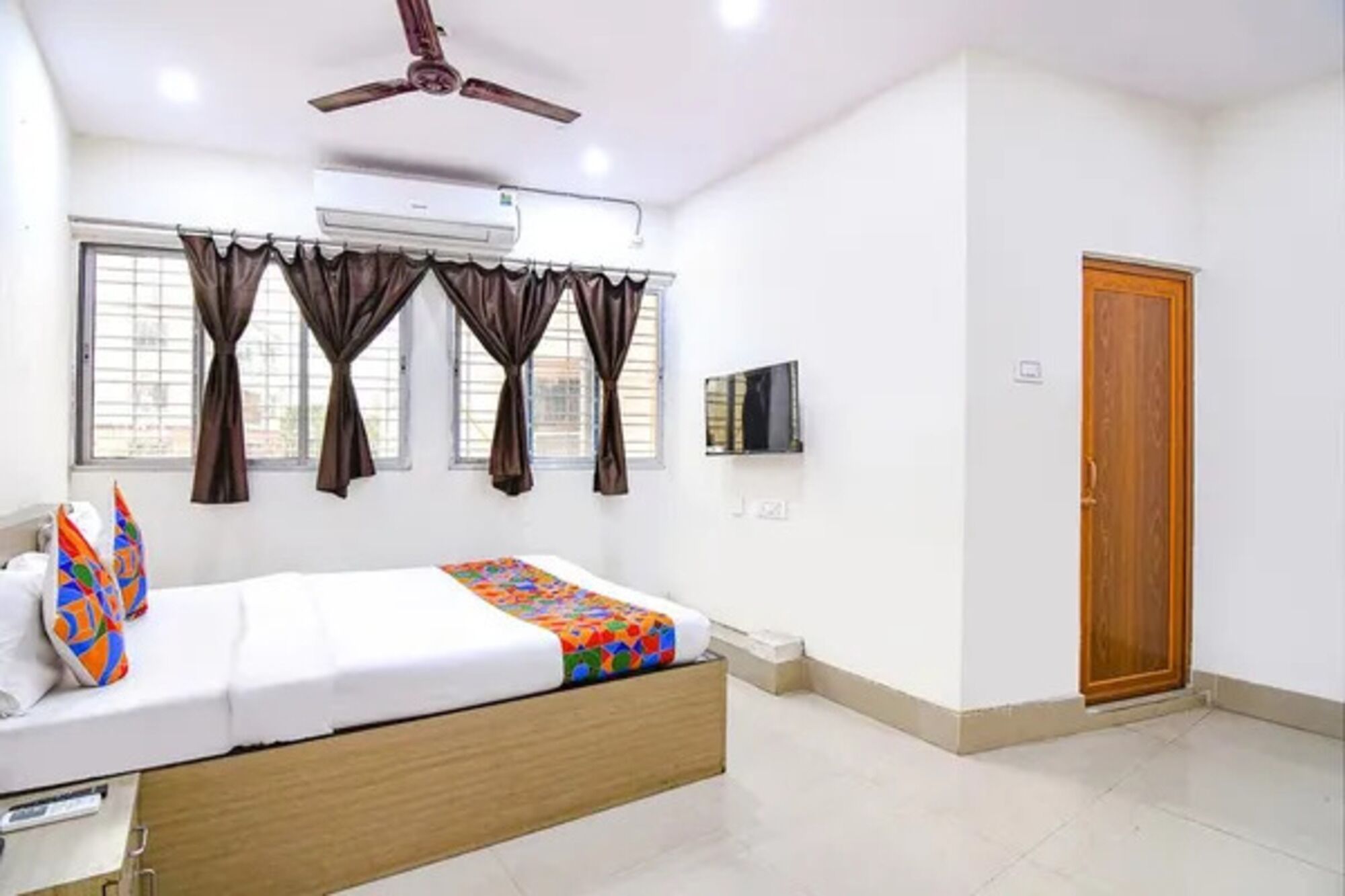 A bedroom with a bed, curtains, a ceiling fan, an air conditioner, a television, and a wooden door.