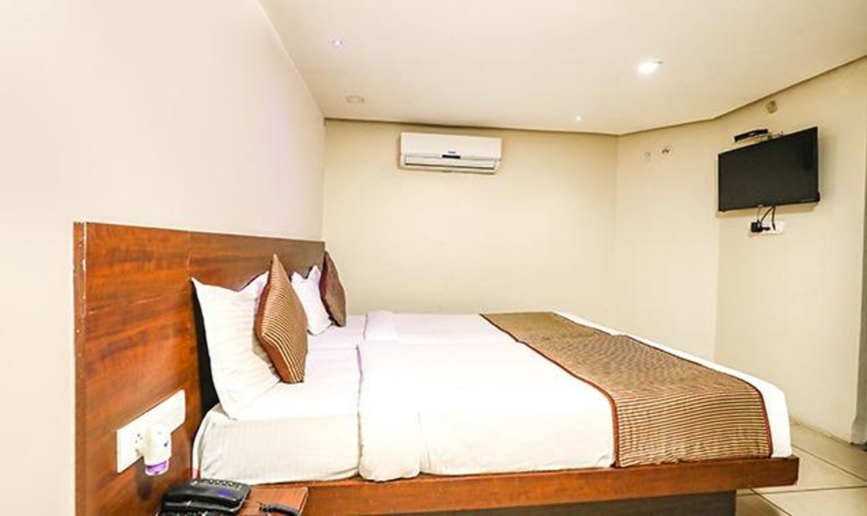 Habitación de hotel con cabecera de madera, televisor de pantalla plana montado en la pared y cama con ropa blanca y una manta marrón.