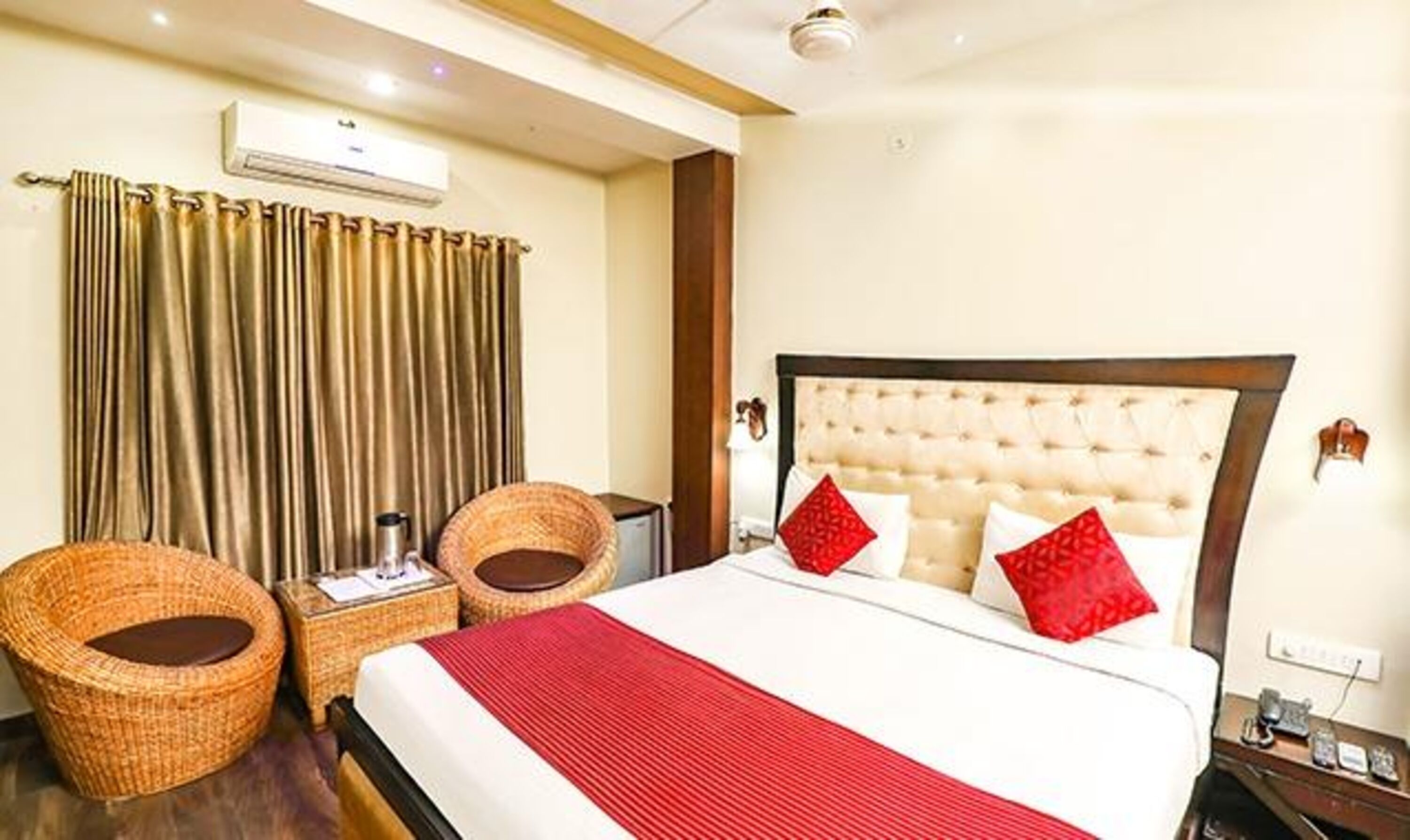A hotel room with a large bed, two wicker chairs, a bedside table with a lamp, and a wall-mounted air conditioner.