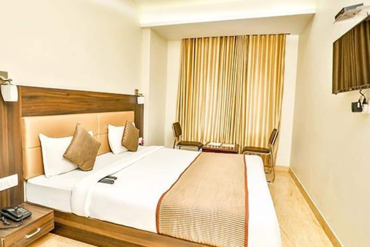 A hotel room with a bed, bedside table, lamp, and a view of the outside through curtains.