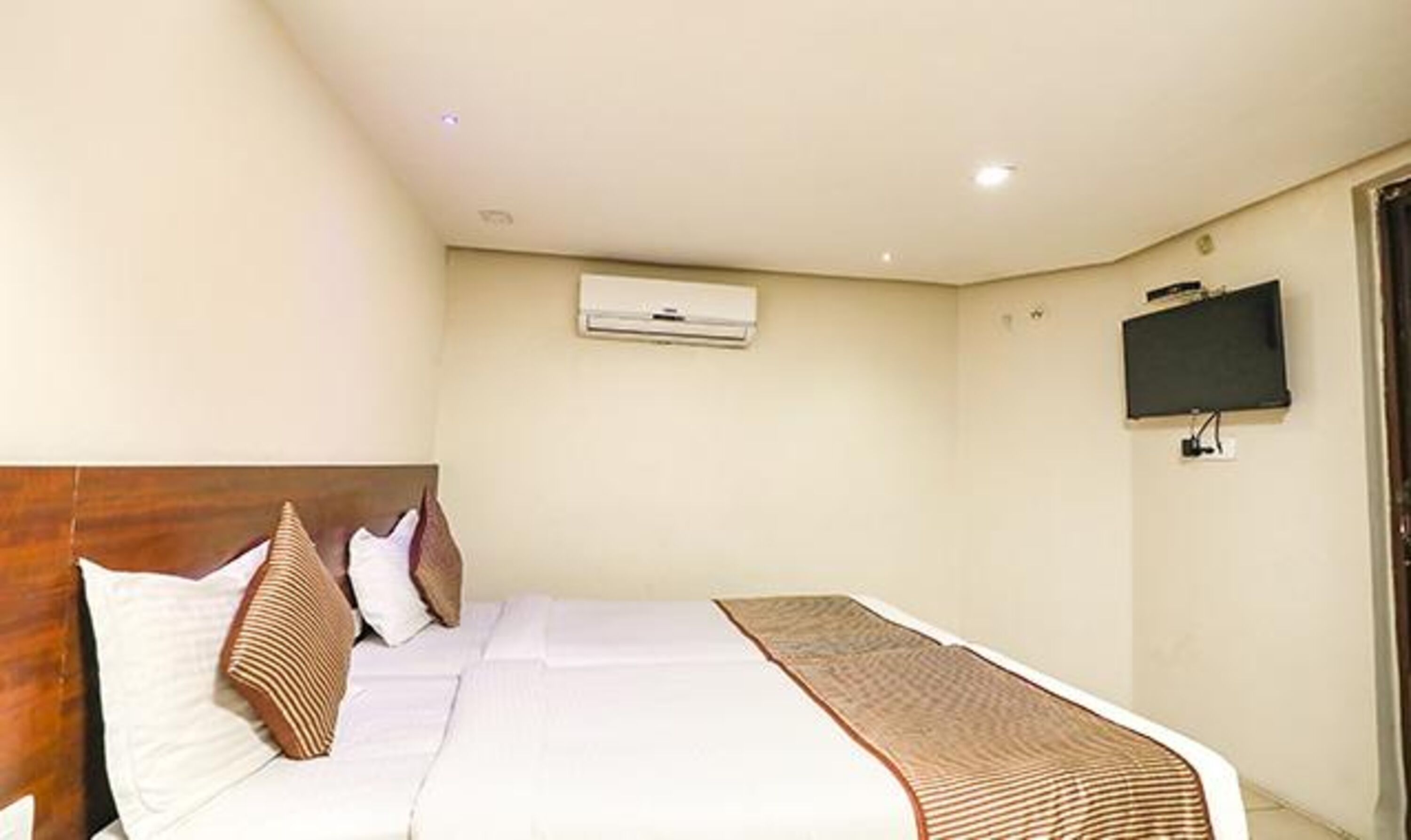 A hotel room with a bed, a flat-screen TV mounted on the wall, and an air conditioning unit above the bed.