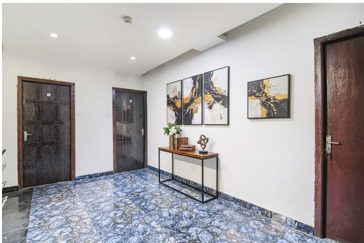 A hallway with patterned flooring, two wooden doors, a console table with decorative items, and three abstract paintings on the wall.