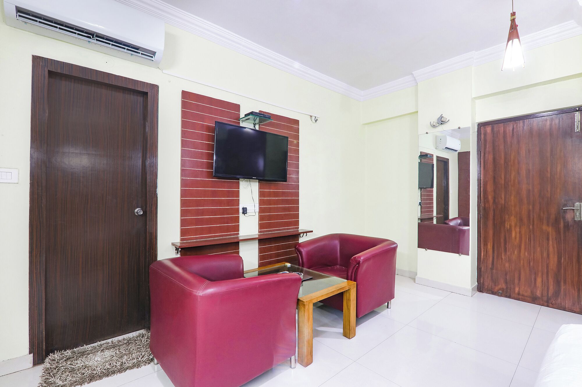 A room with a TV mounted on a brick wall, two red chairs, a small wooden table, and a door with a mirror.