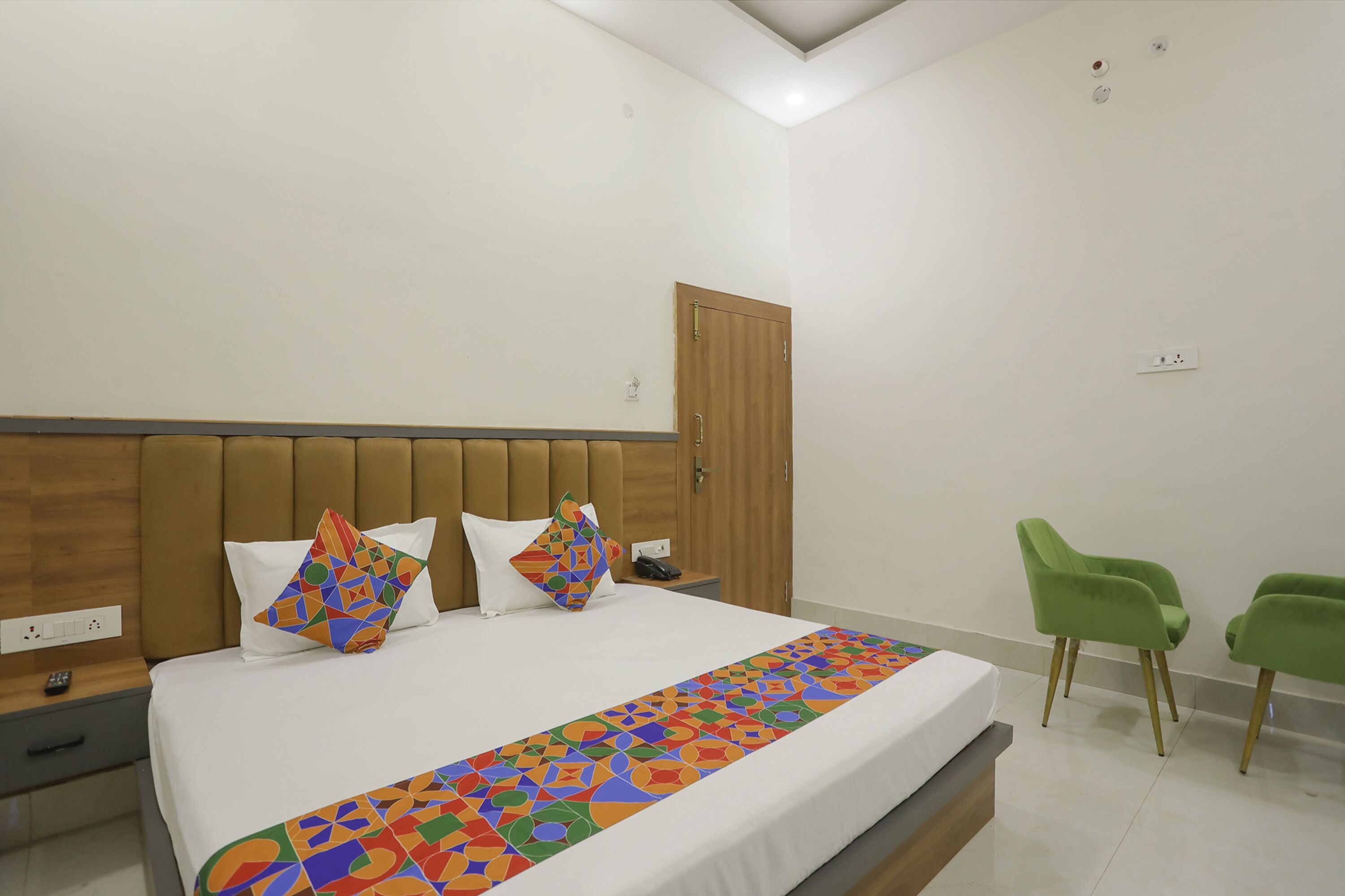 A hotel room with a bed, a wooden headboard, a green chair, and a nightstand with a phone.