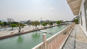 Uma passarela à beira do rio com bancos e árvores, céu claro e um edifício com varanda.
