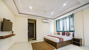 Quarto com cama, ventoinha no teto, ar condicionado, televisão em prateleira, janela com cortinas e uma pequena mesa de cabeceira.