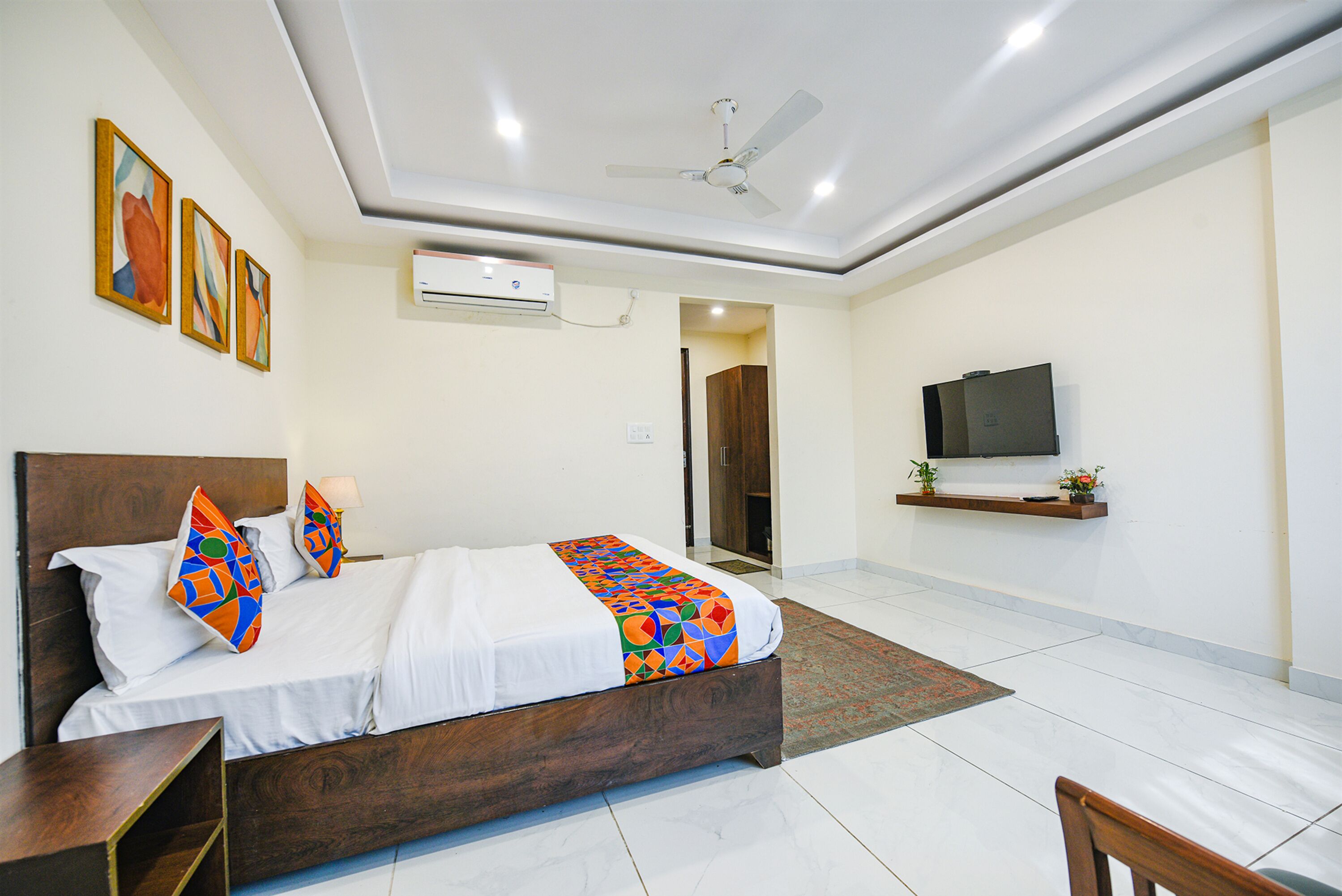 A modern hotel room with a wooden bed, white bedding, colorful pillows, a ceiling fan, an air conditioning unit, a flat-screen TV, and a small shelf with decorative items.
