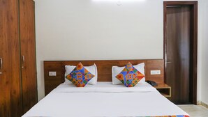 A hotel room with a wooden headboard, a bed with white bedding, two colorful geometric-patterned pillows, a bedside table with a phone, and a wooden wardrobe. - FabHotel Star Corporate Suite I (Gurugram)