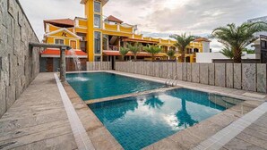 A swimming pool with a wooden deck, a stone wall, and a yellow building with balconies in the background. - Fabhotel Prime Ar Phalazzo Resort (Vanur)