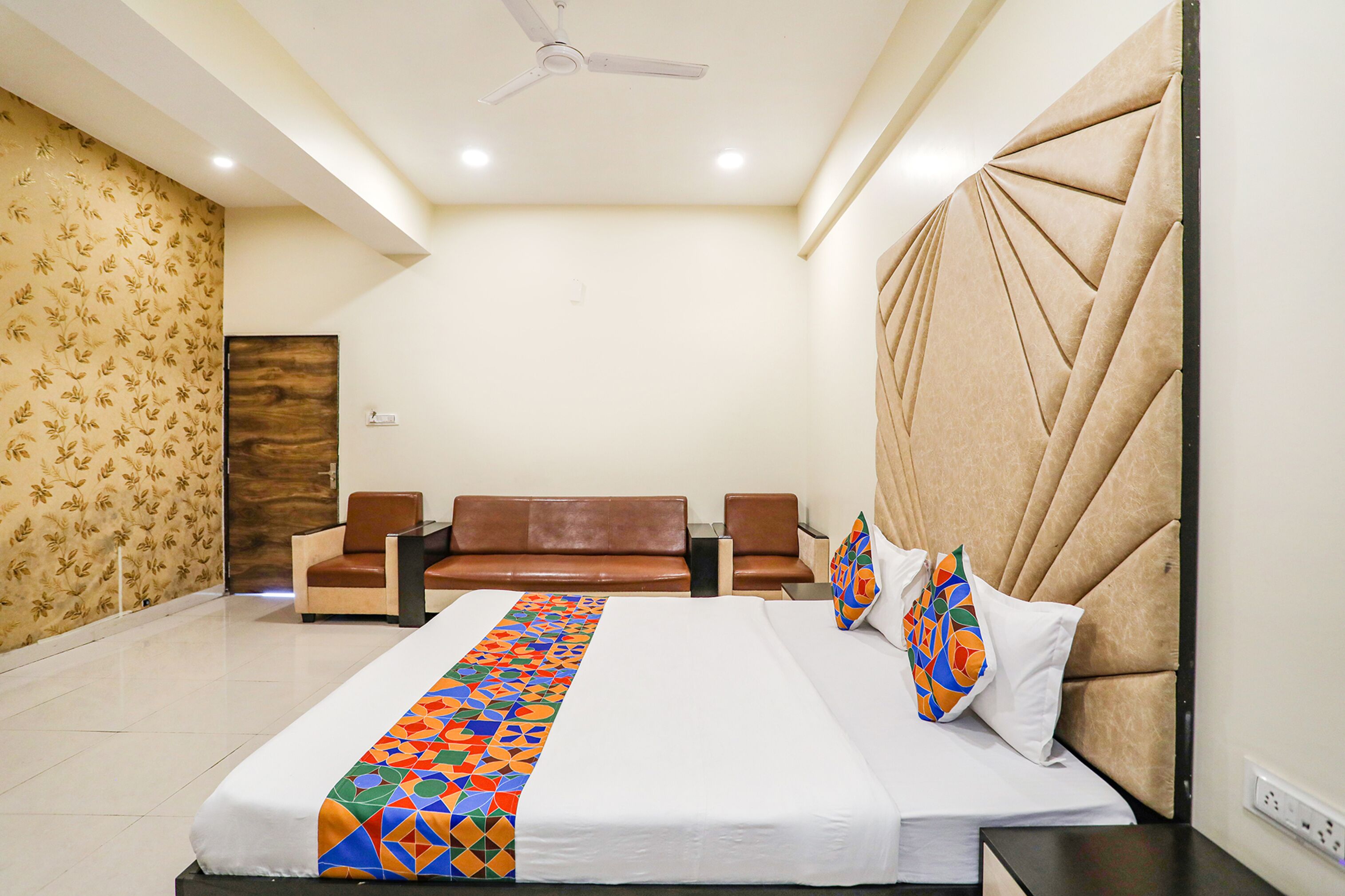A hotel room with a large bed, two brown leather chairs, a ceiling fan, and a patterned wallpaper on one wall.
