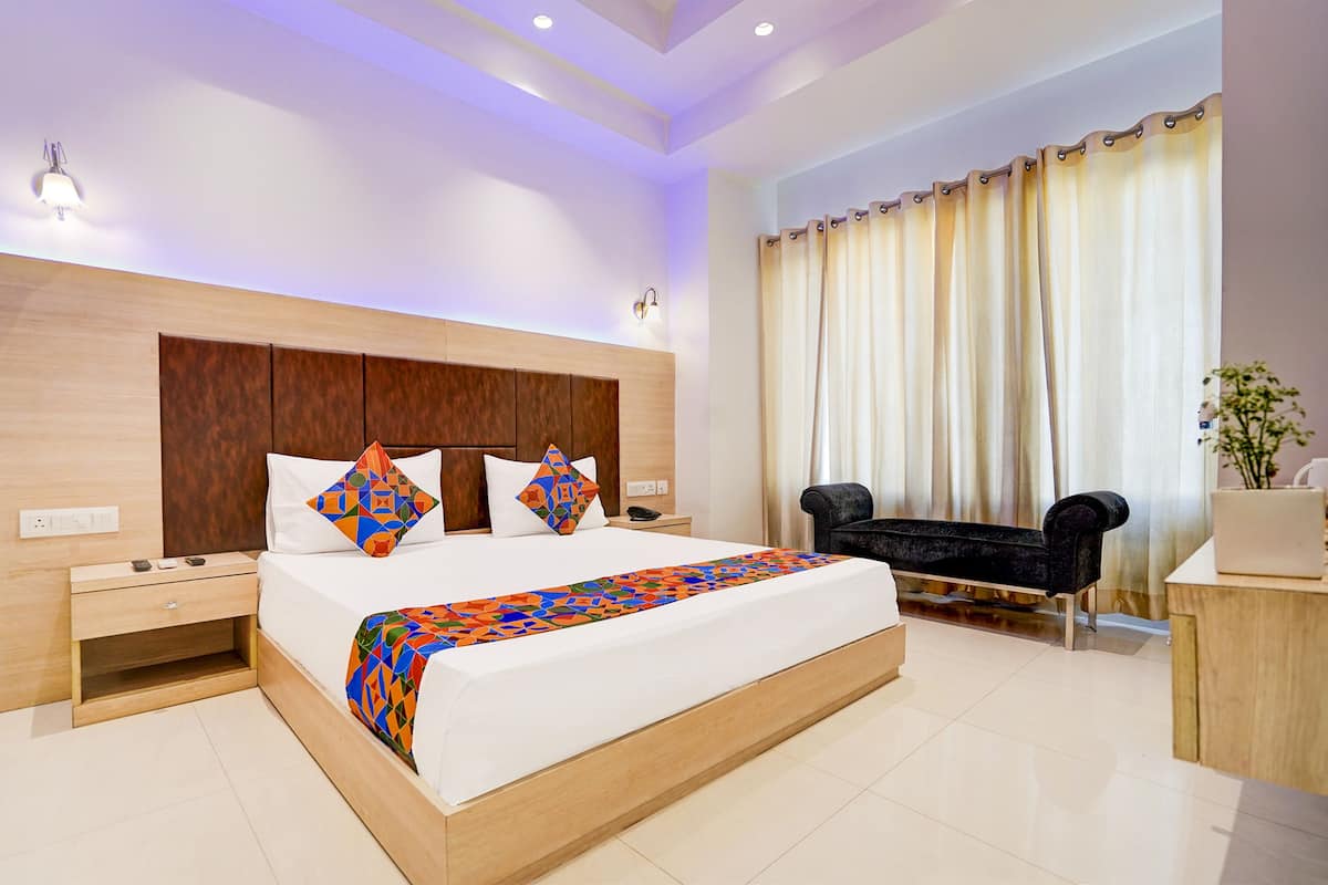 A modern hotel room with a large bed, a wooden headboard, a bedside table, a black sofa, and a large window with curtains.