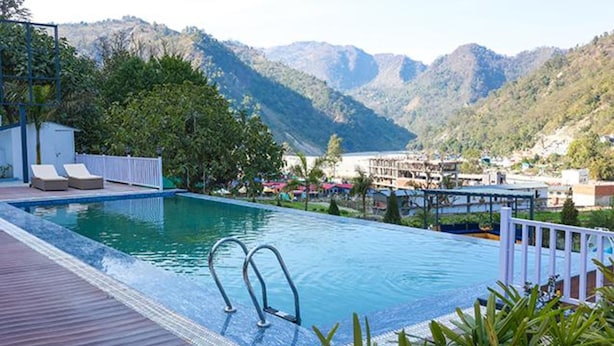 A swimming pool with two lounge chairs and a deck, surrounded by mountains and a view of a resort area. - Fabhotel Prime The Aravali Resort (Yamkeshwar)