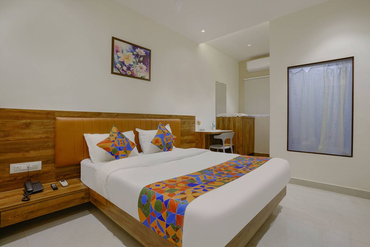 A hotel room with a large bed, a wooden headboard, a bedside table with a phone and remote, a desk with a chair, and a large window with curtains.