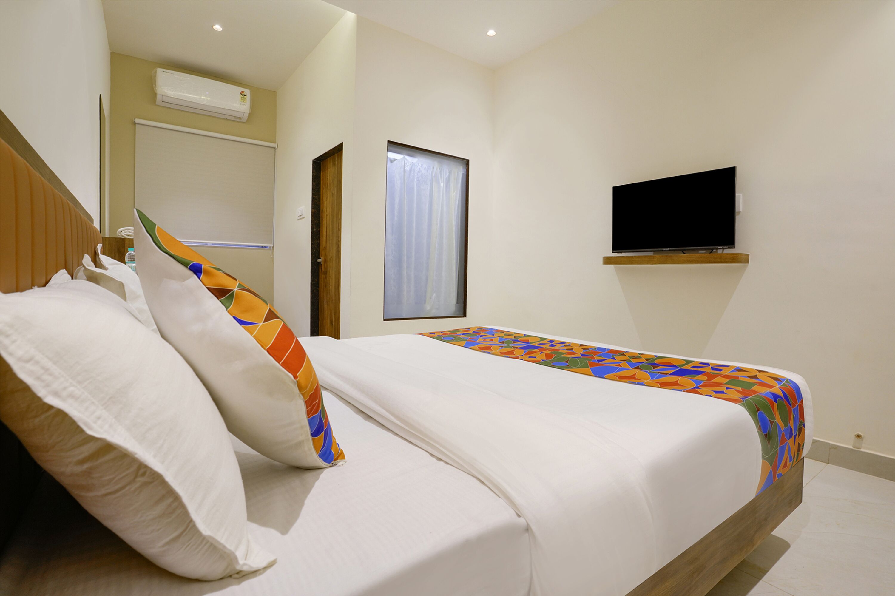 A hotel room with a bed, pillows, a TV on the wall, and a window with curtains.