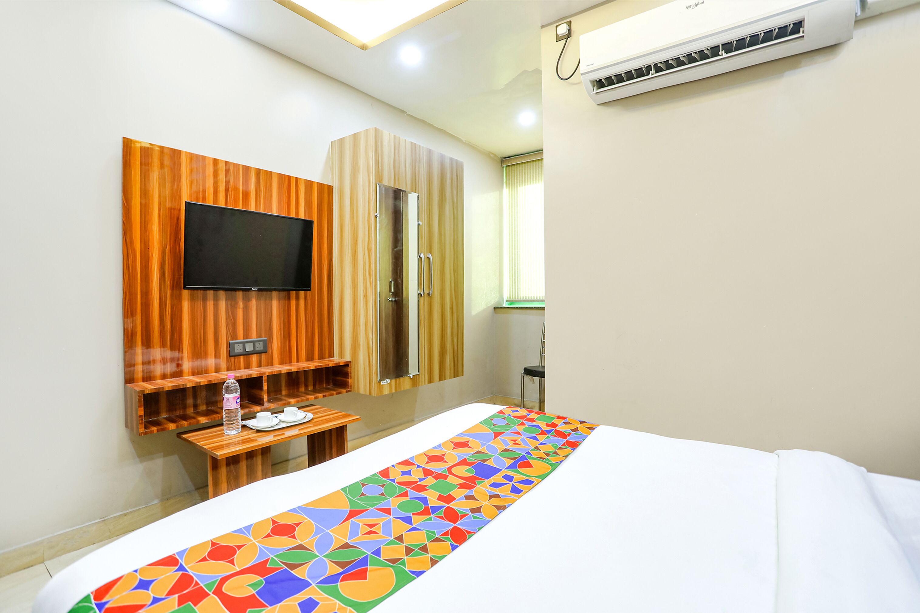 A hotel room with a bed, a TV mounted on a wooden panel, a small table with a bottle and cups, and an air conditioning unit on the wall.