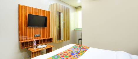 A hotel room with a bed, a TV mounted on a wooden panel, a small table with a bottle and cups, and an air conditioning unit on the wall.