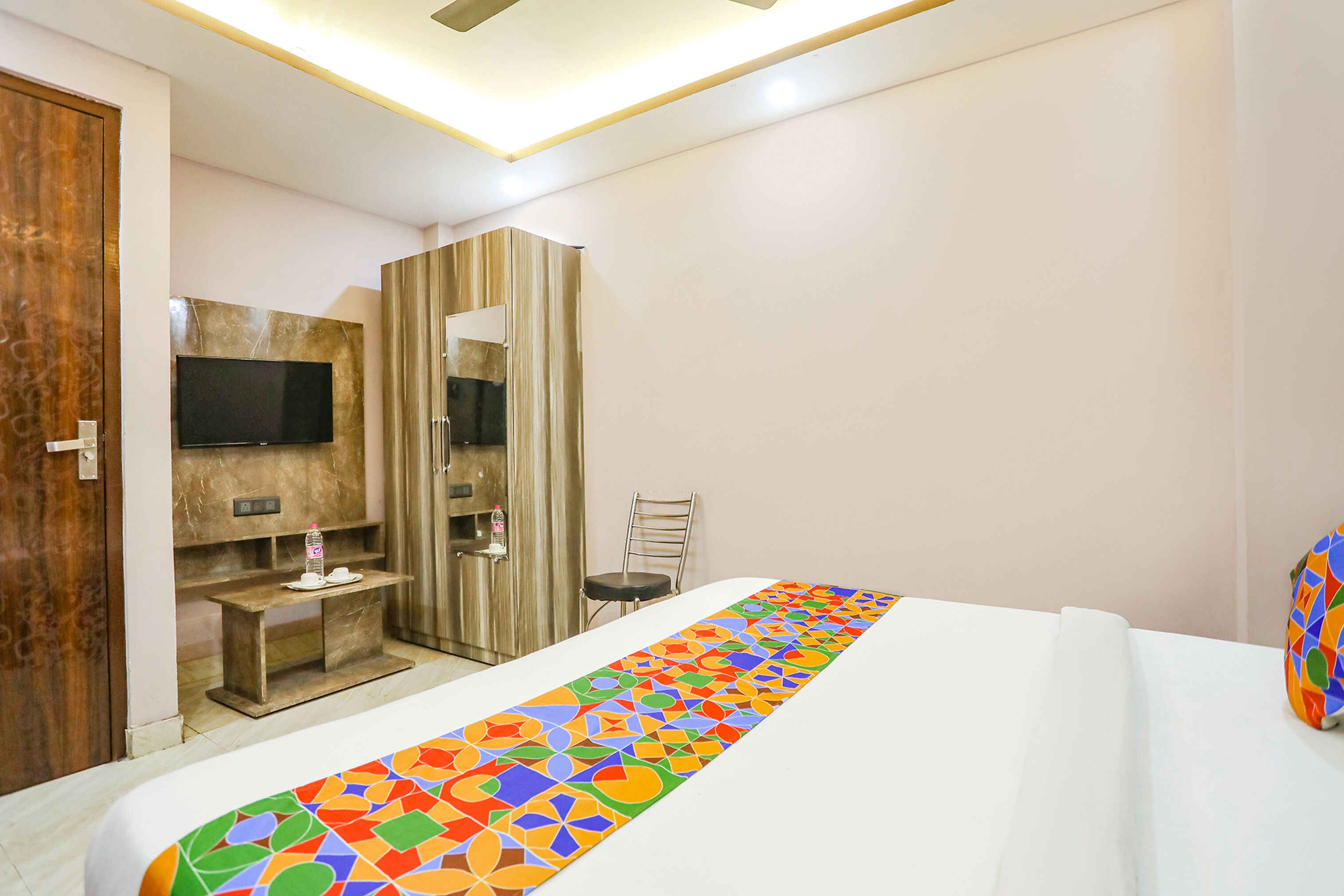 A hotel room with a bed, a TV mounted on a wooden panel, a desk with a chair, and a wardrobe.