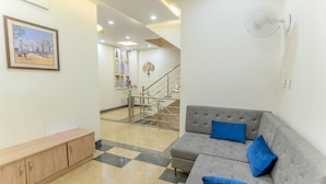 A modern living room with a grey sofa and blue cushions, a wooden cabinet, and a staircase leading to an upper floor. - FabHotel Ramana (Udaipur)