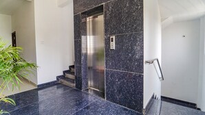 Un ascensor moderno con acabado metálico, junto a una escalera con barandilla y una planta en maceta a la izquierda.