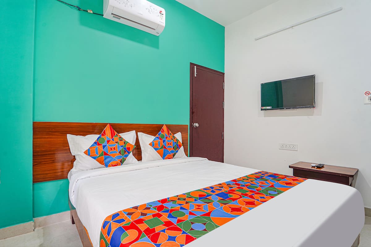 A hotel room with a bed, a wooden headboard, a teal-colored wall, a white air conditioner, a brown door, a small wooden nightstand with a remote control, and a television on the wall.