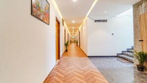 A hallway with a wooden herringbone floor, white walls, and a painting of a cityscape. There are potted plants along the hallway, and a staircase with a stone wall on the right. - Fabhotel Easy Nest (Bengaluru)