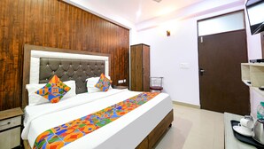 A neatly arranged bedroom with a tufted headboard, a bed with colorful bedding, a wooden wardrobe, a small table with a kettle, and a door. - Fabexpress Golden Stone (Ghaziabad)