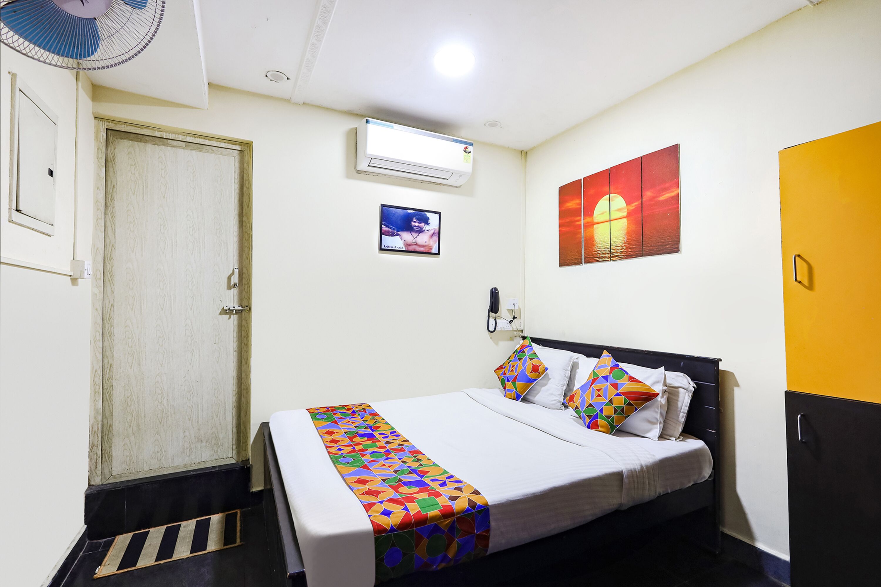 Cama bem arrumada com um edredom colorido de padrão geométrico, um ar condicionado fixado na parede, uma moldura com uma foto de pôr do sol e um telefone na parede.
