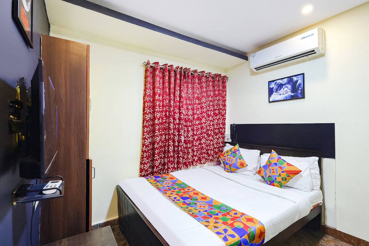 A hotel room with a bed, a TV, an air conditioner, and a window with red curtains.