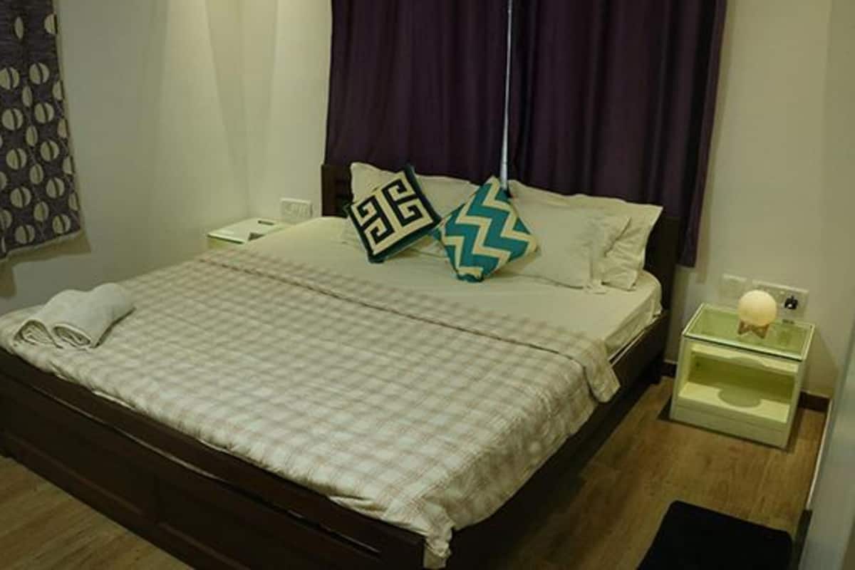 Un lit bien fait, recouvert d’une couvre-lit à carreaux, deux oreillers aux motifs géométriques, une table de chevet avec une lampe et un mur agrémenté d’un rideau.
