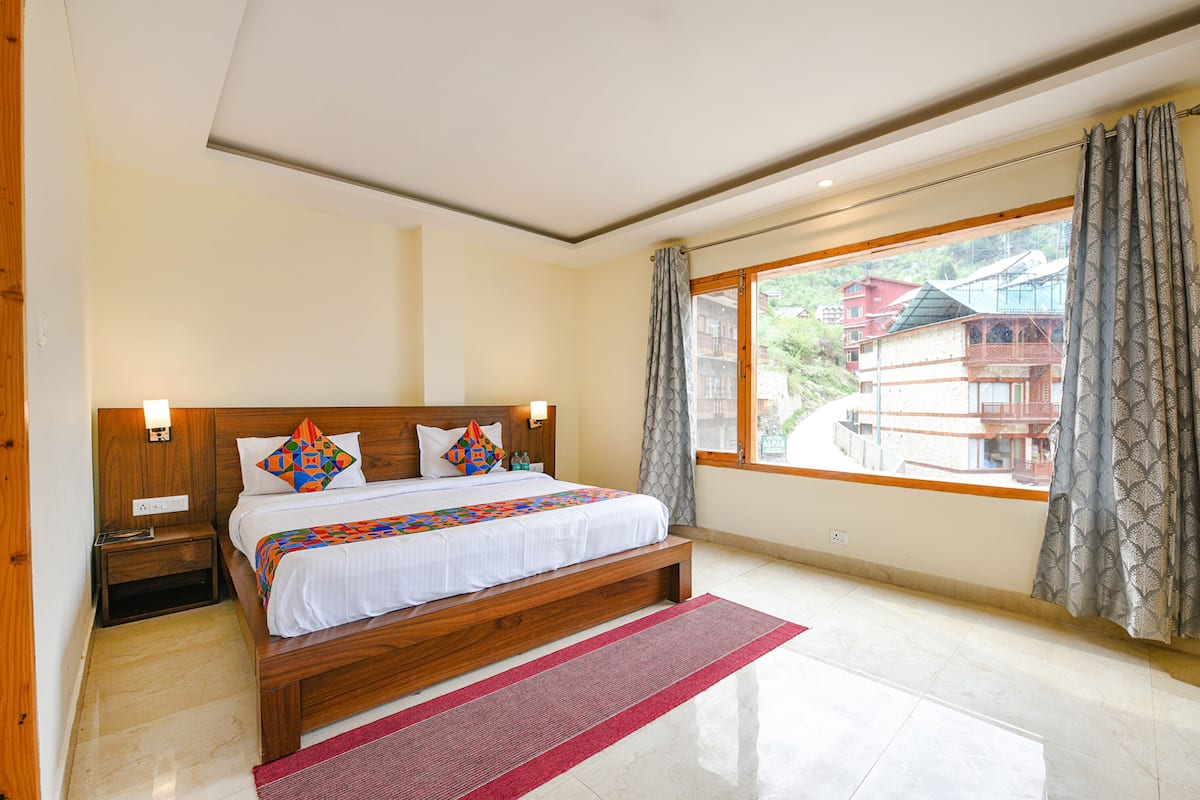 A bedroom with a wooden bed, a nightstand, a window with curtains, and a view of buildings outside.