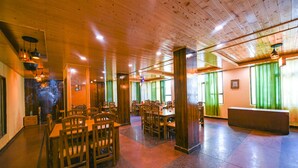 A spacious dining area with wooden tables and chairs, a stone wall, and green curtains on the windows. - Fabhotel Prime The Kalinga Grand (Manali)