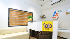 Een receptiebalie met een 'fab hotels' bord, een receptiegebied met een raam bestaande uit houten latten, een receptiebalie met een 'receptie' aanduiding, en een logo voor 'Ajinkya Group' aan de muur.