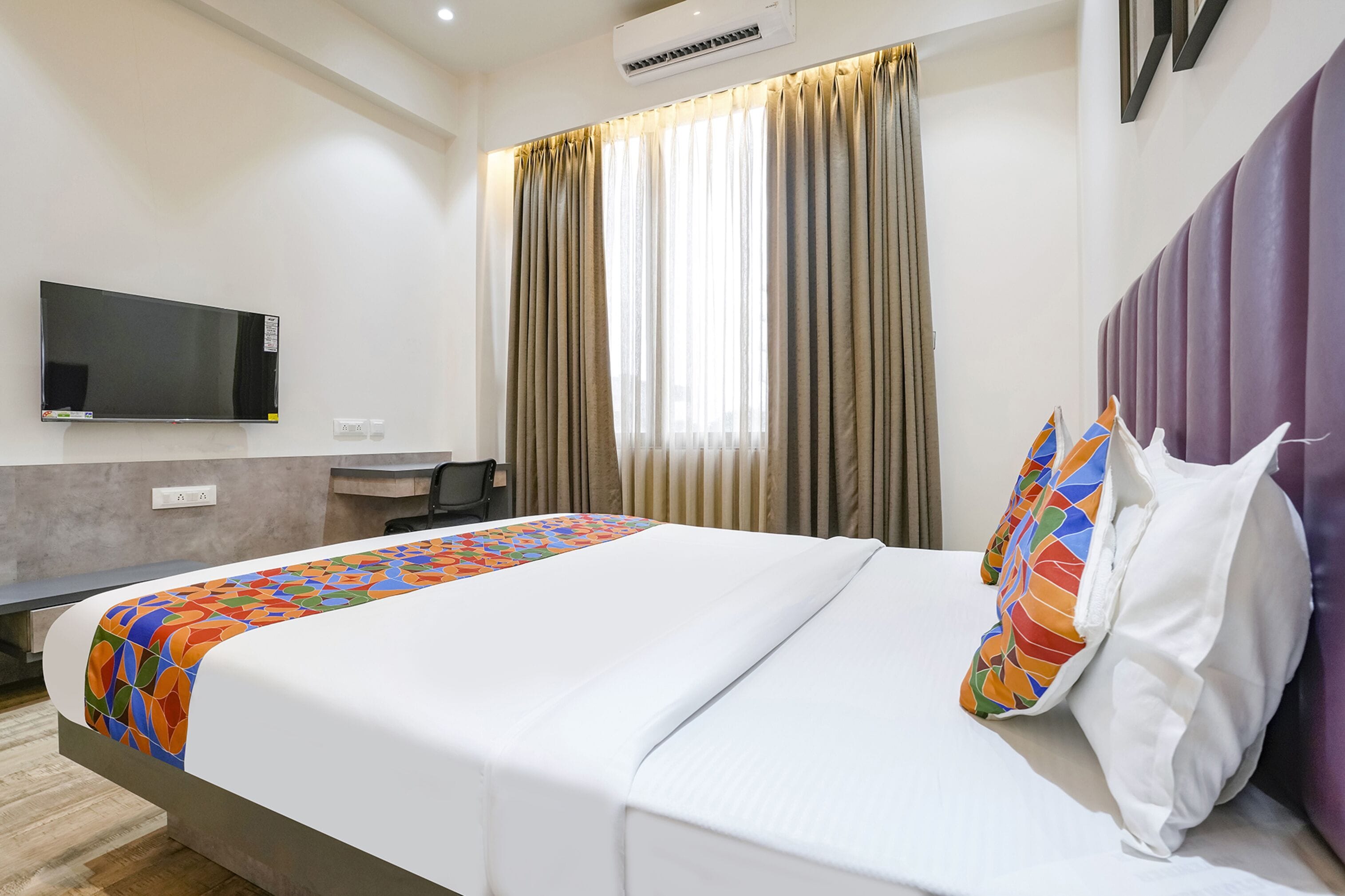 A hotel room with a bed, a flat-screen TV mounted on the wall, a desk, and a window with curtains.