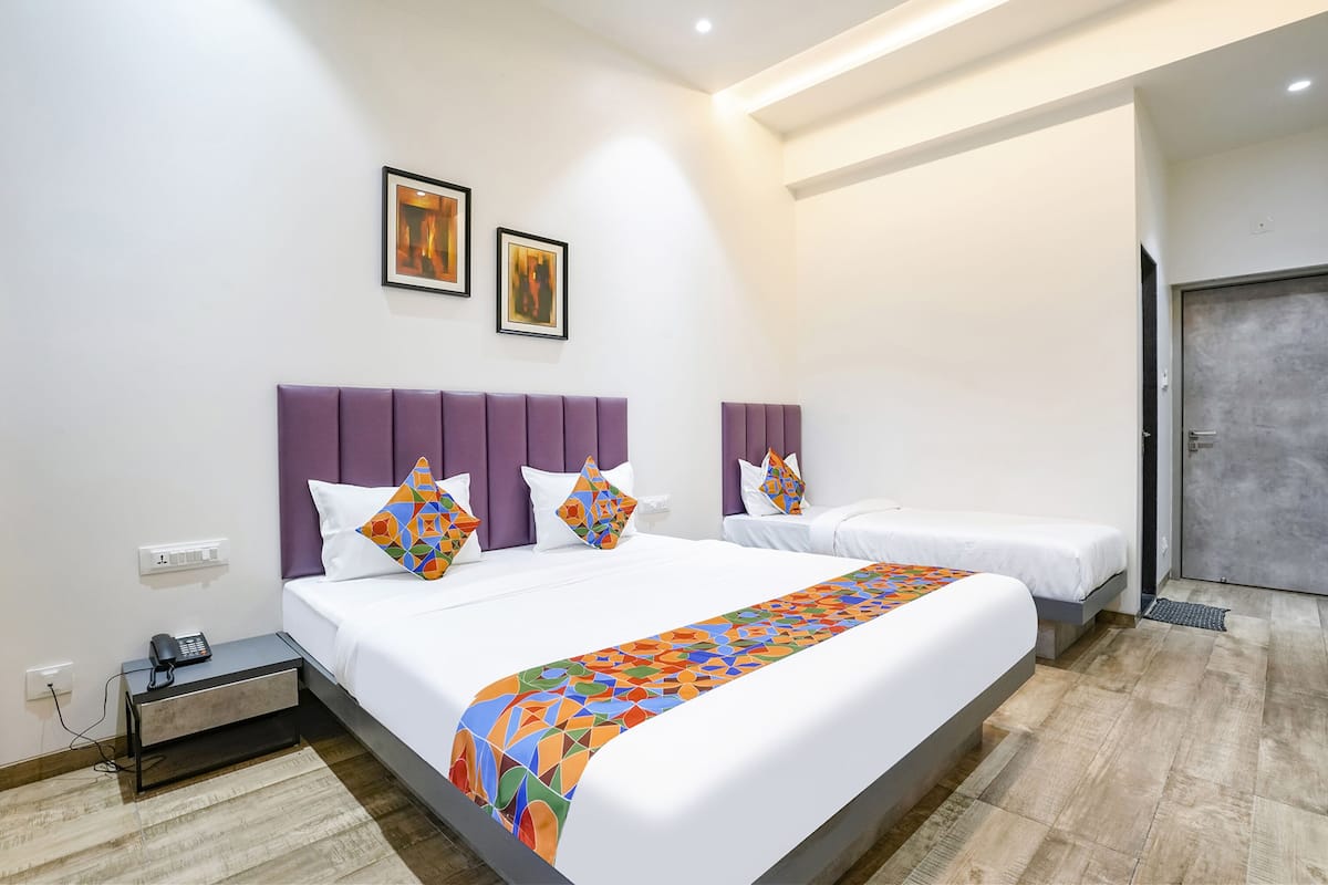 Ein Hotelzimmer mit zwei Betten, jeweils mit violetten Kopfteilen, weißer Bettwäsche und farbenfrohen, geometrisch gemusterten Tagesdecken. An der Wand hängen zwei gerahmte Bilder, auf dem Nachttisch steht ein Telefon und rechts ist eine Tür zu sehen.