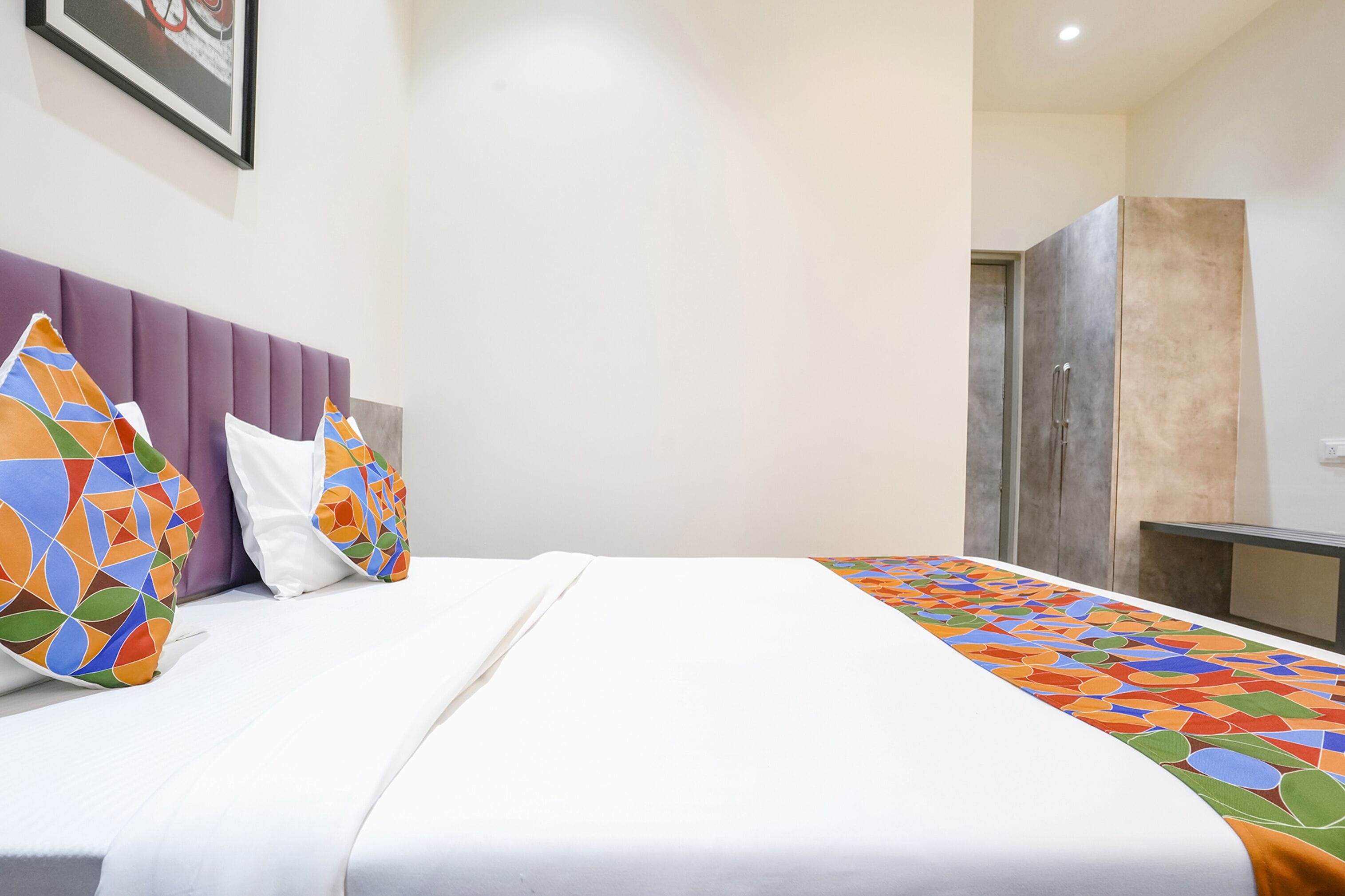 A hotel room with a bed, a headboard, a wardrobe, and a framed picture on the wall.