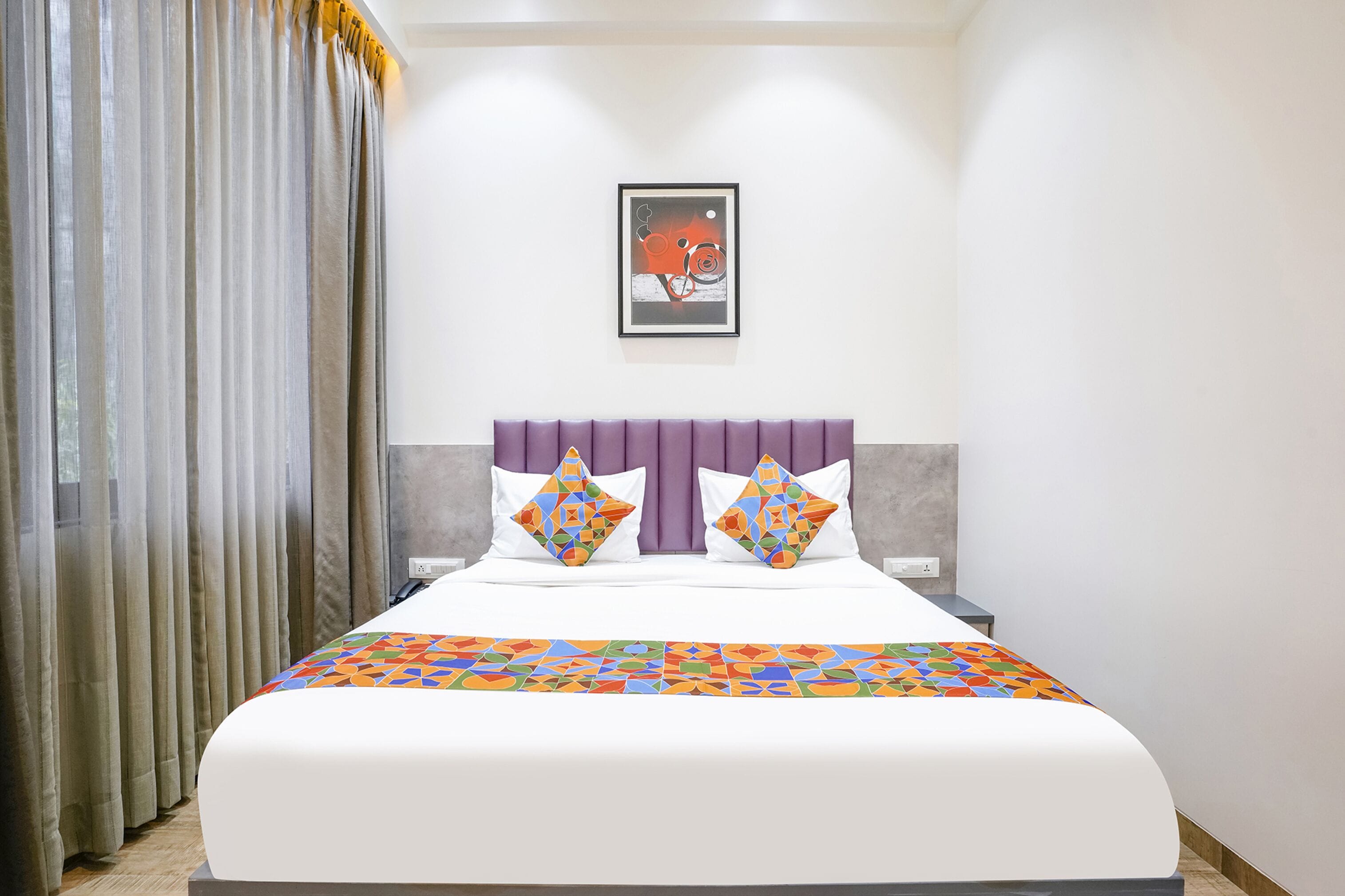A modern hotel room with a large bed, a purple headboard, and a colorful geometric-patterned bedspread.