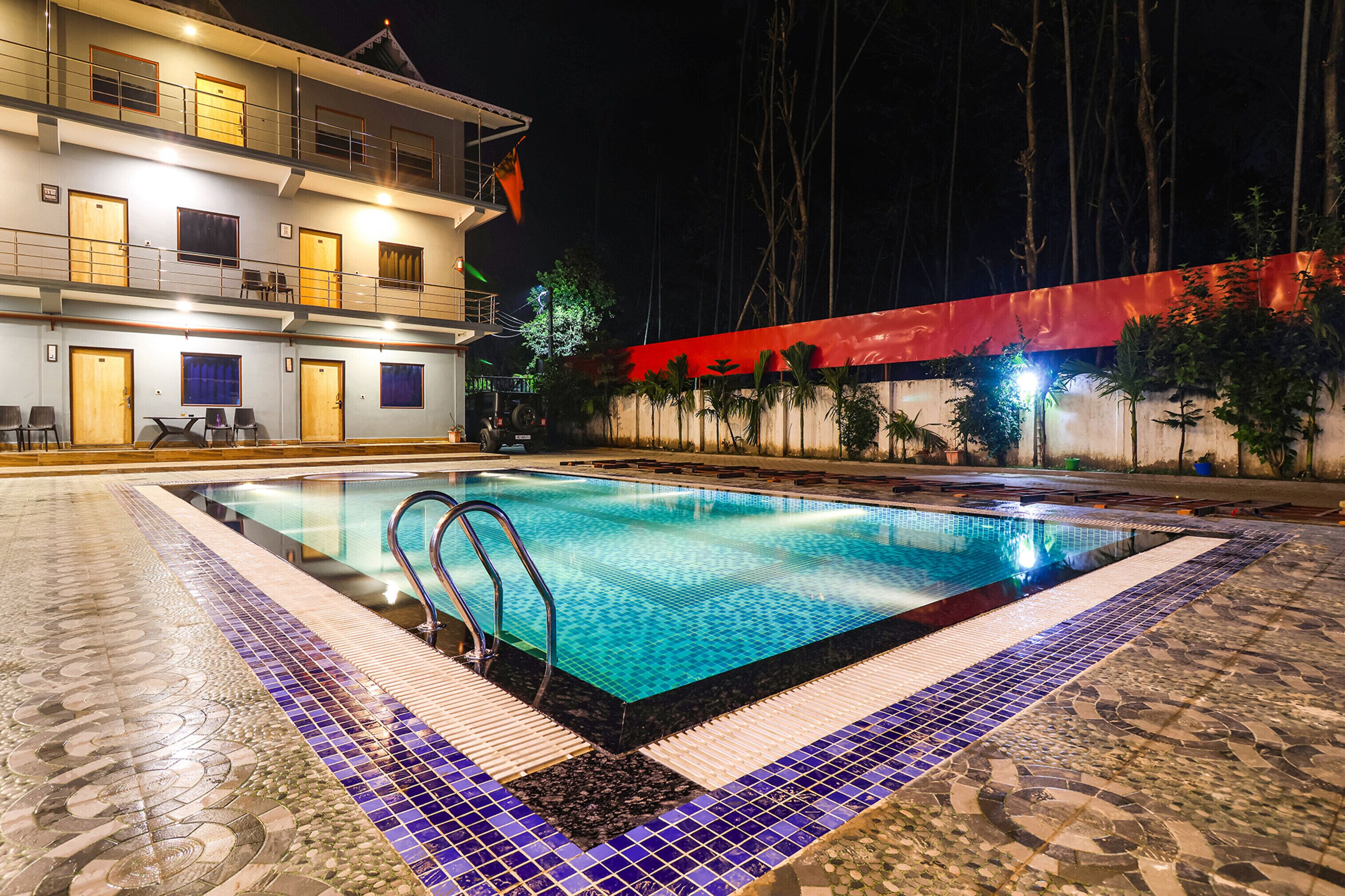 A swimming pool with a tiled deck, a building with multiple balconies, and a red wall with plants.