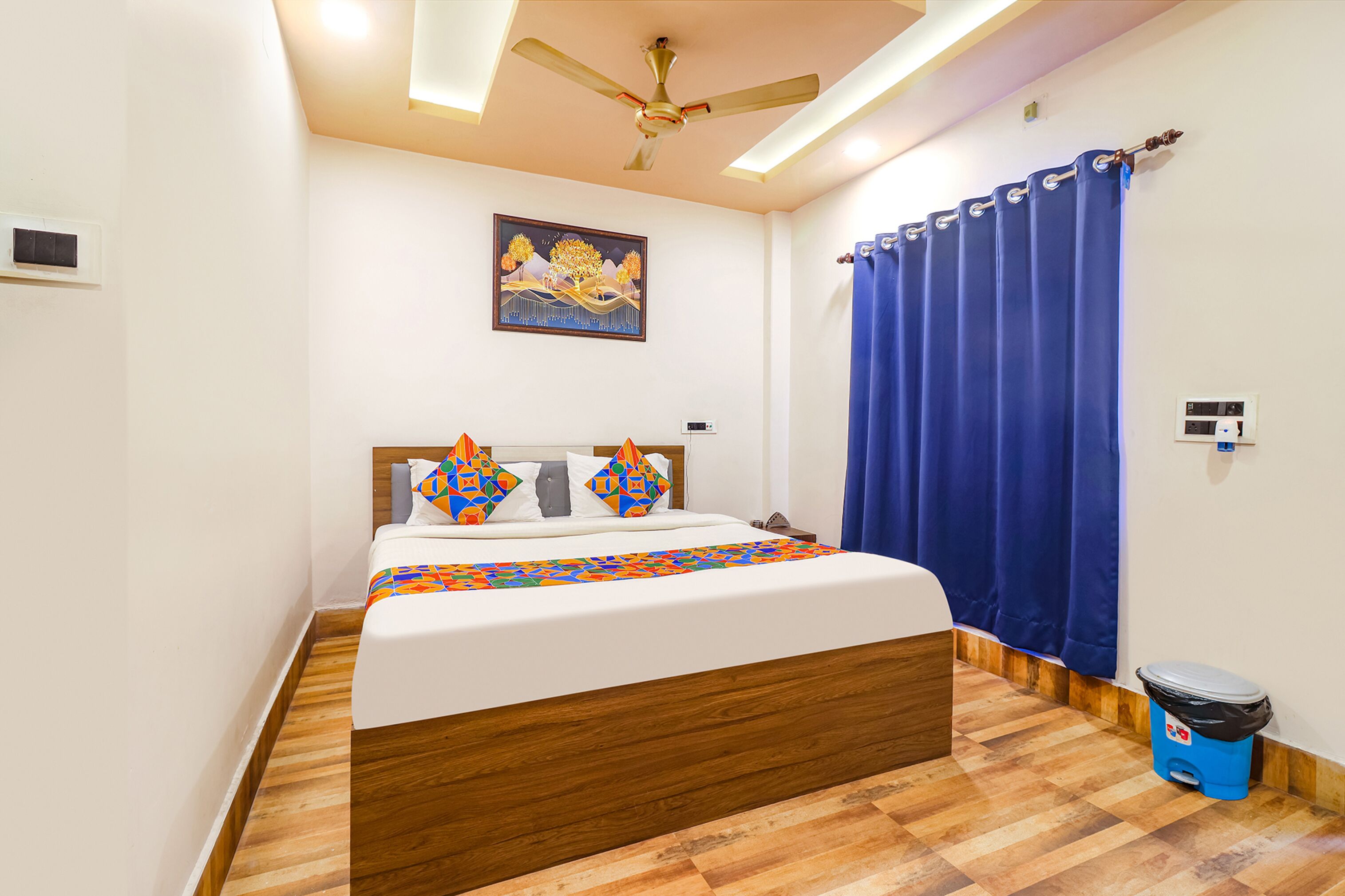 A bedroom with a bed, a ceiling fan, a painting, a blue curtain, and a trash bin.